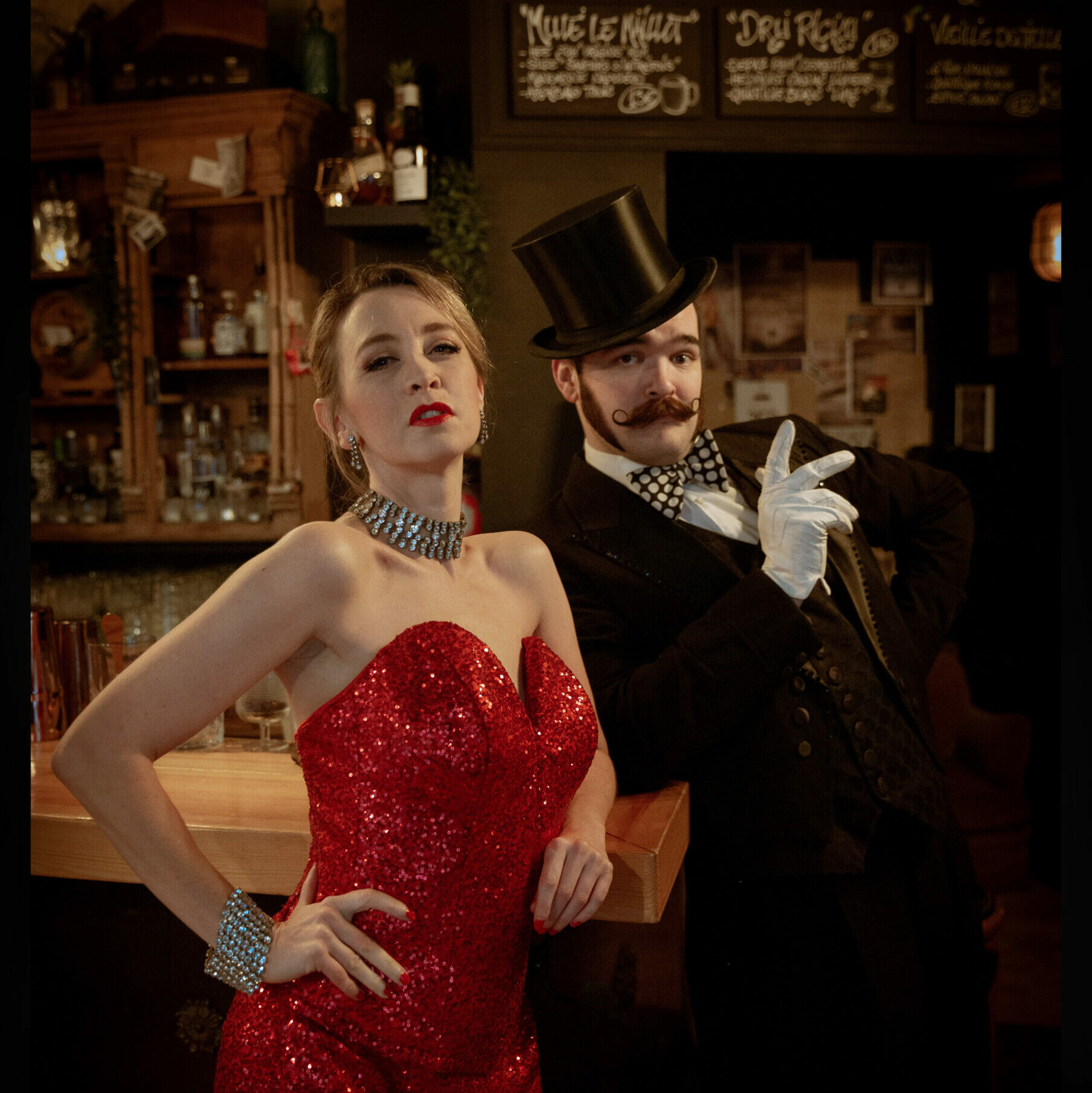 Affiche du spectacle Boudoir Burlesque par Betty Crispy et Russell Bruner. On voit les deux artistes devant un bar. Betty Crispy porte une robe rouge pleine de paillette. Russell Bruner quant à lui porte un chapeau haut de forme, un costard noir et un noeud papillon.